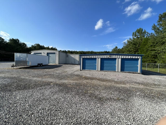 Small storage building with three blue roll-up doors beside a larger metal building